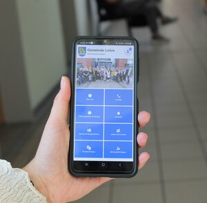 Bild vergr&ouml;&szlig;ern: Eine Frau hat ein Smartphone in der Hand, auf dem die Startseite der App "Gemeinde Lehre" zu sehen ist.
