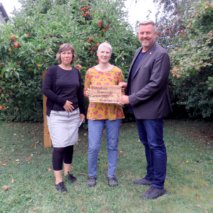 Bild vergr&ouml;&szlig;ern: Gartenbesitzerin Karin Noodt bekam von Bürgermeister Andreas Busch und Verwaltungsmitarbeiterin Eike Guhl neben dem Preisgeld auch ein Holzschild geschenkt. Dieses erinnert an Ihre Teilnahme am Gemeindewettbewerb naturnahe Gärten.