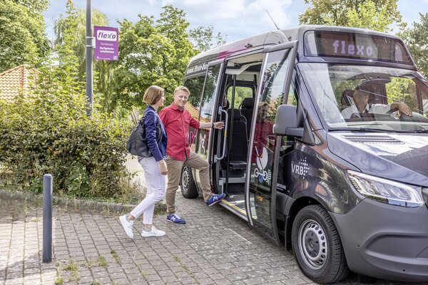 Ein Flexobus hält an einer Haltestelle und eine männliche und eine weibliche Person möchten dort einsteigen.