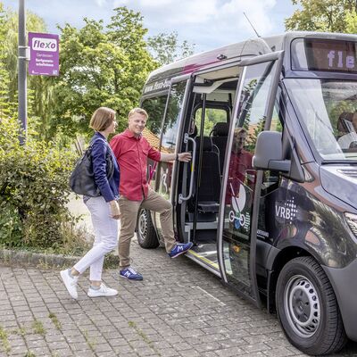 Ein Flexobus hält an einer Haltestelle und eine männliche und eine weibliche Person möchten dort einsteigen.