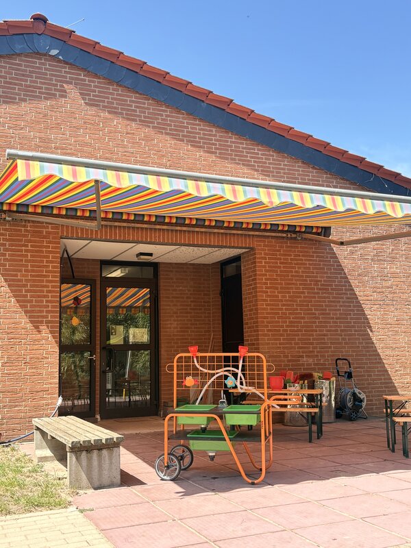 Bild vergr&ouml;&szlig;ern: Die Terrasse ist mit einer getreiften Markise in Regenbogenfarben überspannt und darunetr stehen Bänke und Spielgeräte.