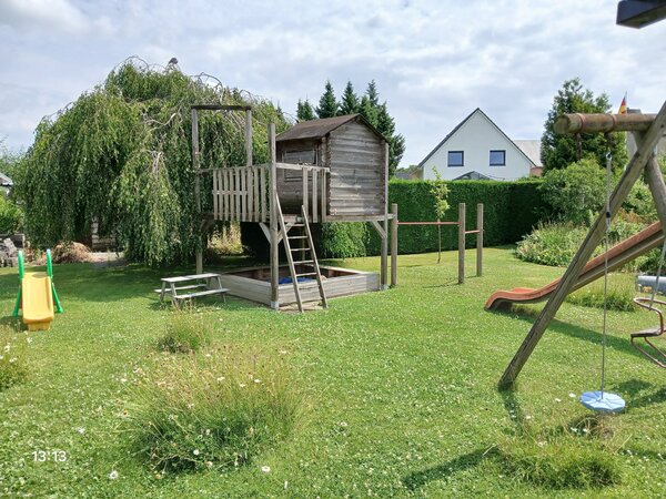 Bild vergr&ouml;&szlig;ern: In einem großen Garten steht eine Rutsche und ein Speilhaus aus Holz. Dort ist noch ein Sandkasten und Turnstangen integriert.