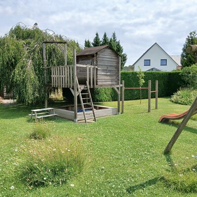 In einem großen Garten steht eine Rutsche und ein Speilhaus aus Holz. Dort ist noch ein Sandkasten und Turnstangen integriert.