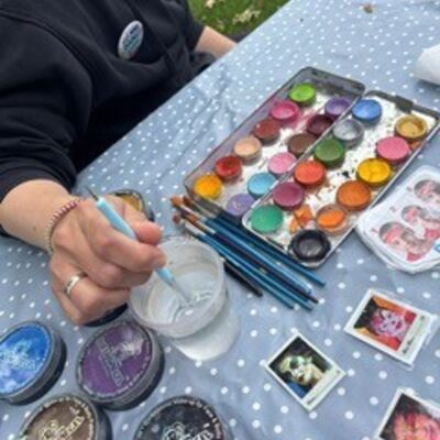Bild vergr&ouml;&szlig;ern: Eine weibliche Person wäscht einen Pinsel in einem Wassergals aus. Daneben leiegn auf dem Tisch eine Schminkpalette mit vielen bunten Farben.