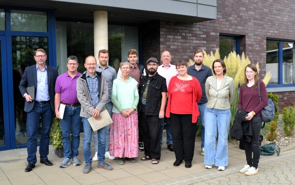 Man sieht eine Gruppe von zwölf Menschen die in zwei Reihen vor einem Gebäude stehen.