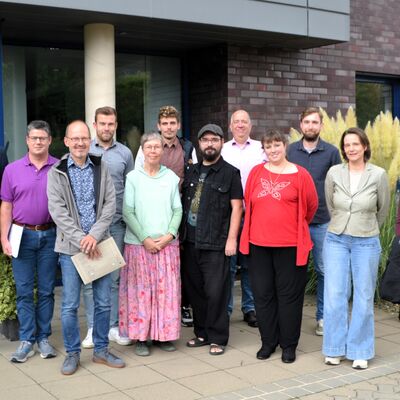 Man sieht eine Gruppe von zwölf Menschen die in zwei Reihen vor einem Gebäude stehen.