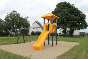 Bild vergr&ouml;&szlig;ern: Ein Spielplatz mit einer Sandfläche im Vordergrund, auf der eine gelbe Rutsche steht mit zwei Schaukeln.