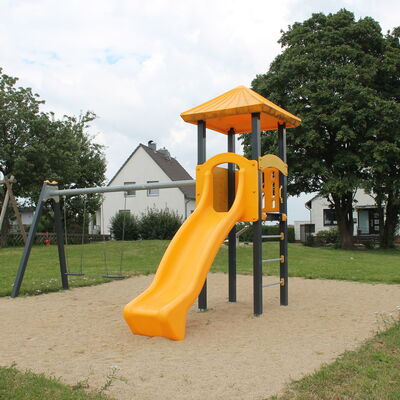 Ein Spielplatz mit einer Sandfläche im Vordergrund, auf der eine gelbe Rutsche steht mit zwei Schaukeln.