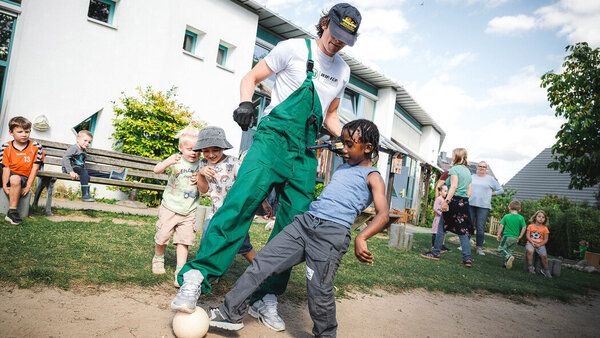 Bild vergr&ouml;&szlig;ern: Ein Profispieler in Arbeitslatzhose soielt mit einem kleinen Jungen Fußball.