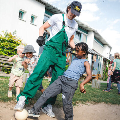 Ein Profispieler in Arbeitslatzhose soielt mit einem kleinen Jungen Fußball.