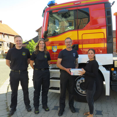 Vier Personen stehetn vor dem Führerhaus eines Feuerwehrfahzeuges. Frau WEstpahl übergibt Herrn Görke eine kleine befüllte durchsichtige Plastikbox.