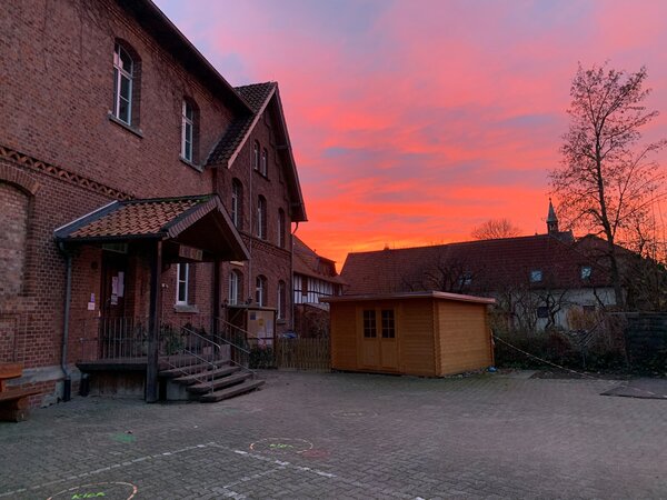 Bild vergr&ouml;&szlig;ern: Man sieht den Eingang und ein Teil des Hofes der Kita, wobei der Himmel rosa-orange vom Licht des Sonnenuntergangs gefärbt ist.