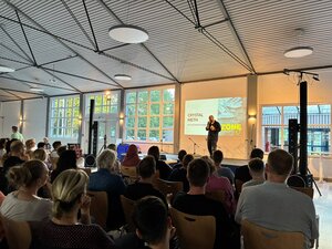 Bild vergr&ouml;&szlig;ern: Auf der Bühne steht Podcaster Sebastian Caspar, auf der Wand hinter ihm ist eine Präsentation projeziert. Im Vordegrund sieht man den gefüllten Zuschaueraum in der Börnekenhalle.
