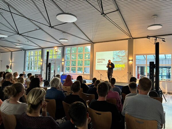 Auf der Bühne steht Podcaster Sebastian Caspar, auf der Wand hinter ihm ist eine Präsentation projeziert. Im Vordegrund sieht man den gefüllten Zuschaueraum in der Börnekenhalle.
