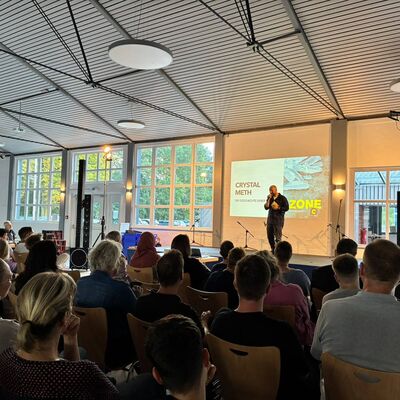 Auf der Bühne steht Podcaster Sebastian Caspar, auf der Wand hinter ihm ist eine Präsentation projeziert. Im Vordegrund sieht man den gefüllten Zuschaueraum in der Börnekenhalle.