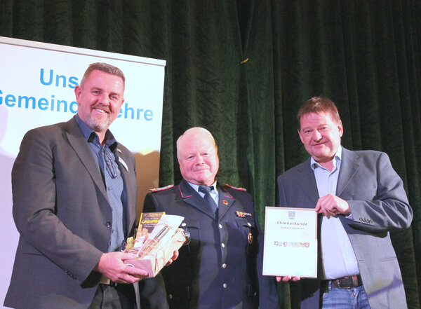 Bild vergr&ouml;&szlig;ern: Drei Männer stehen vor einem dunklen Vorhang. In der Mitte steht der Geehrte in Feuerwehrunifom. Linkst steht der Bürgermeister, der ein Präsent überreicht. Rechts von ihm übergibt der Ortsbürgermeister die Urkunde.