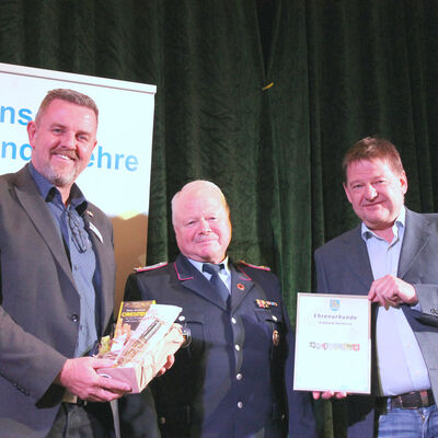Drei Männer stehen vor einem dunklen Vorhang. In der Mitte steht der Geehrte in Feuerwehrunifom. Linkst steht der Bürgermeister, der ein Präsent überreicht. Rechts von ihm übergibt der Ortsbürgermeister die Urkunde.
