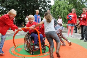 Bild vergr&ouml;&szlig;ern: Auf einem inklusiven Spielplatz: Ein Junge im Rollstuhl wird von drei anderen Kindern auf einem Karussell gedreht.