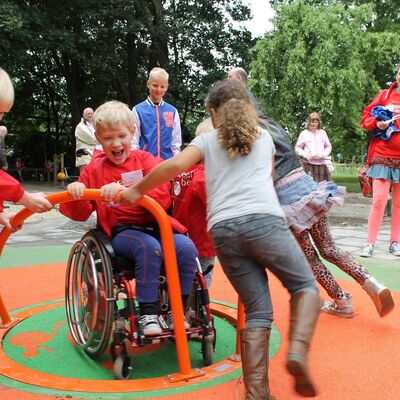 Auf einem inklusiven Spielplatz: Ein Junge im Rollstuhl wird von drei anderen Kindern auf einem Karussell gedreht. 